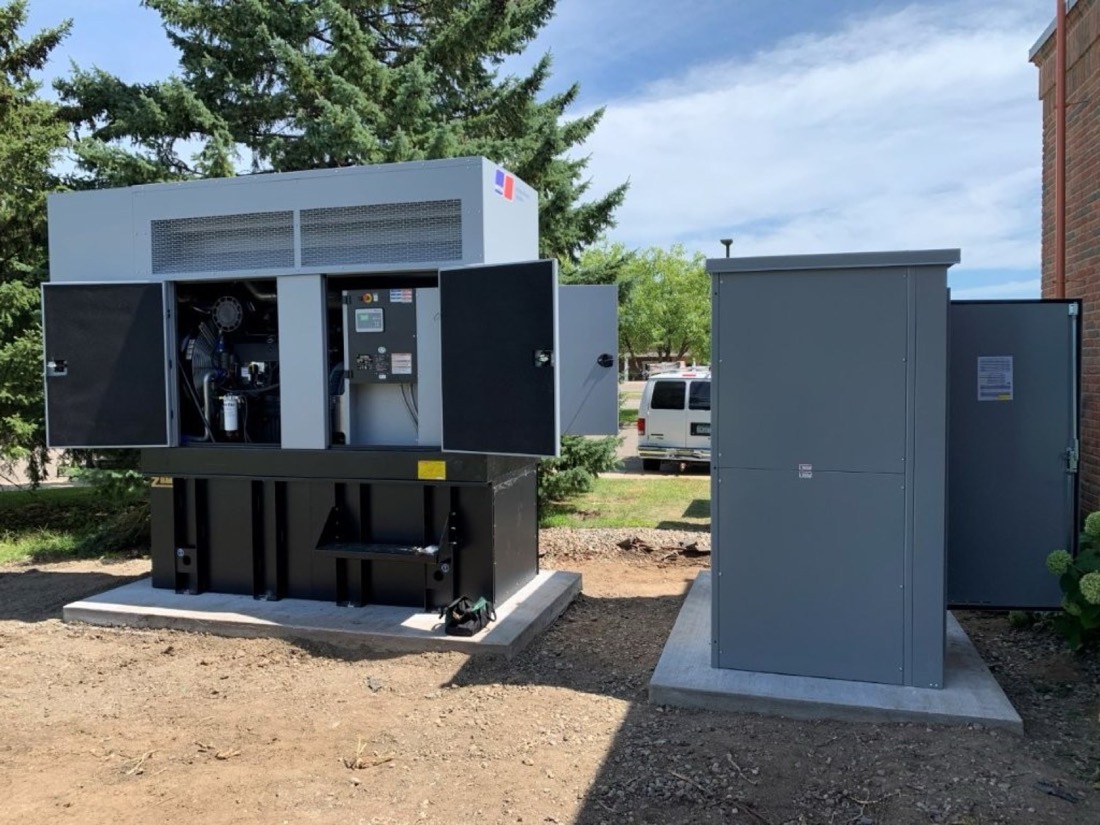A standby generator with its enclosure doors open, beside outdoor electrical cabinets, both set on new concrete pads.